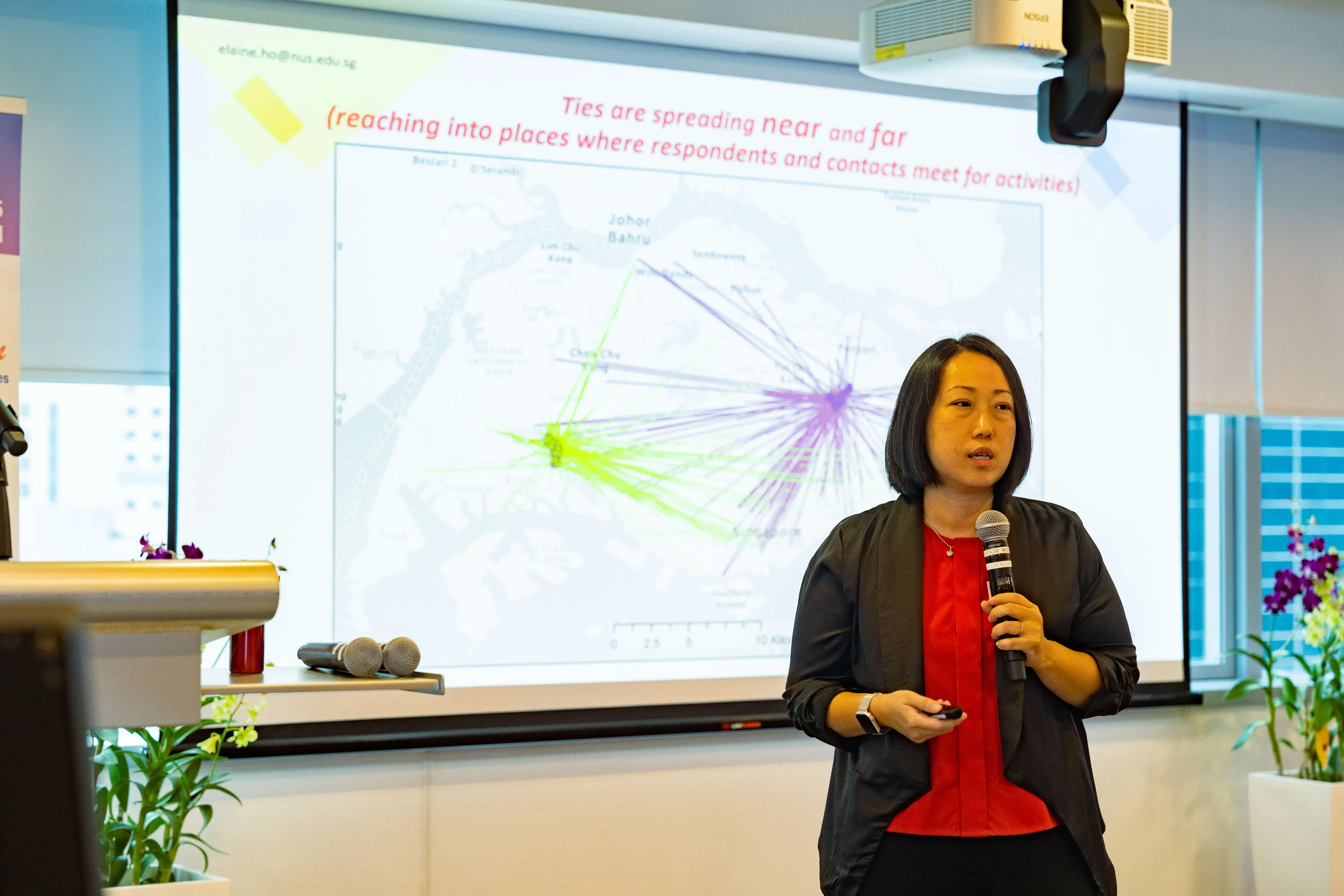 Woman in red top and gray blazer speaks into a microphone in front of a projected map slide about social ties.